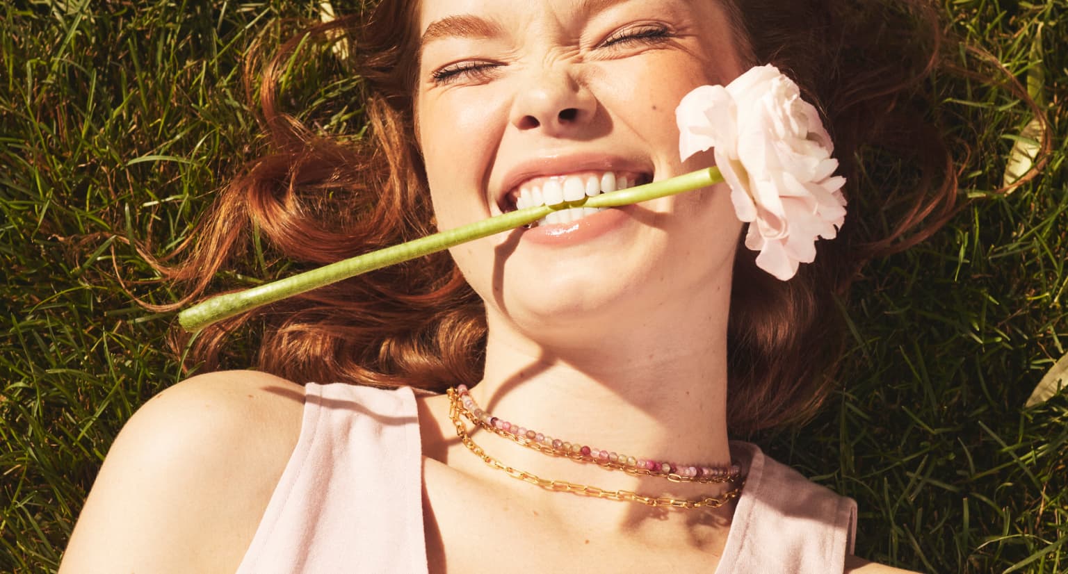 a woman laying in grass holding a white flower in her teeth